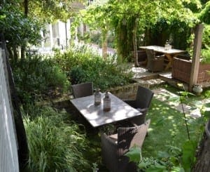 A small garden features a square table with four wicker chairs, surrounded by lush greenery and shaded by trees. Mirrors placed strategically among the plants reflect the vibrant scenery. A second table is visible in the background.