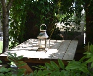 A metal lantern sits on a wooden picnic table surrounded by green plants, trees, and decorative mirrors in an inviting outdoor garden setting.