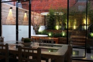 A modern dining area in a stylish building features a glass wall that overlooks a landscaped patio garden with plants, wooden fencing, and outdoor lighting.