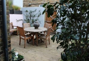 Wooden patio furniture set with a round table and four chairs sits on a wooden deck. A large ceramic vase is on the table, surrounded by lush planting design and a wooden fence.