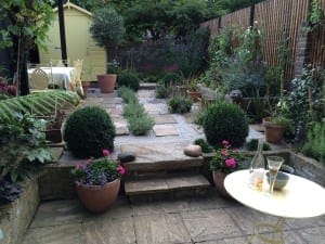 A small garden design with stone paths, potted plants, trimmed bushes, a yellow shed, and a round table set with wine glasses and a bottle.