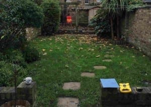A small backyard with green grass, scattered leaves, and signs of modern family life: a red toy swing, a mug, a blue paper, and a yellow toy resting on a low brick wall.