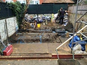 A person stands in a backyard with dug foundation trenches, building tools, bags of debris, and materials scattered around the construction site.