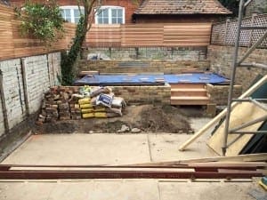 A backyard building site under construction with bricks, cement bags, and dirt piles; a partially built brick wall and wooden planks are visible in the background.