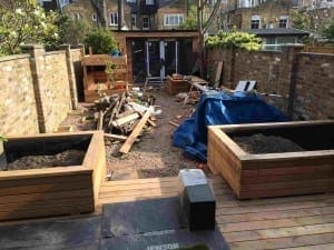 A backyard under renovation for a modern family, featuring wooden planters, scattered construction materials, a pile of wood, and a blue tarp covering part of the ground.