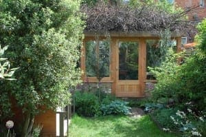 A wooden garden shed with large glass windows and a door, surrounded by green bushes, trees, and various plants. Branches rest on the roof, giving the scene the charm of a nature-inspired sketch.
