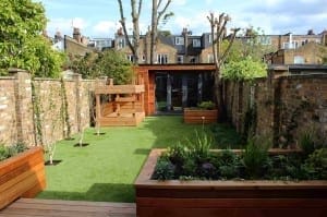 A modern backyard designed for the modern family, featuring artificial grass, raised wooden planters, climbing plants on brick walls, and a wooden garden office at the far end.