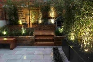 Modern backyard garden with wooden steps, brick walls, planters, and bamboo plants illuminated by ground lights in the evening, showcasing stylish building elements.