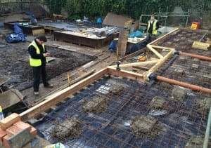 Two construction workers in high-visibility vests, guided by project management principles, prepare a reinforced concrete foundation with steel rebar and wooden formwork on a busy building site.
