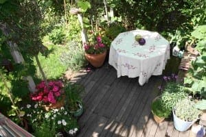 A small wooden patio edible garden with potted plants and flowers, featuring a round table covered with a white floral tablecloth and three decorative bowls.
