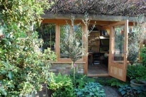 A small garden office with wooden-framed windows and open doors, surrounded by lush green plants, trees, and an edible garden, with books and a desk visible inside.