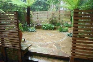 A small garden patio with circular stone paving, wooden slat fences, and lush green plants feels like a green room sanctuary, especially when viewed on a rainy day amid ferns and gentle showers.