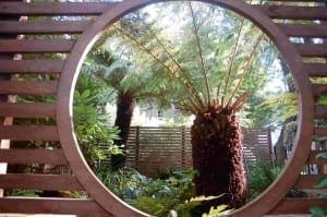 A round wooden frame in a slatted fence creates a green room effect, showcasing a tree fern and lush garden greenery. Sunlight filters through the trees in the background.