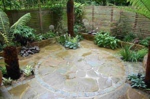 A small garden with stone patio flooring arranged in a circular pattern, surrounded by various green plants, creates a natural green room feeling and is enclosed by wooden fencing.