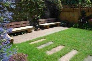 Two wooden benches on a stone patio invite relaxation in this chillout garden, surrounded by green grass, stepping stones, a wooden fence, and blooming flowers.
