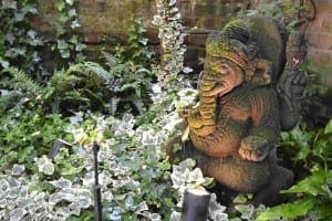 A moss-covered stone statue of Ganesha, radiating Eastern Exotic charm, is surrounded by green foliage and ivy in a shaded garden area.