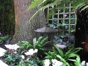 A small stone lantern sits under a tree in an Eastern exotic garden, surrounded by ferns, white flowers, and a wooden lattice fence.