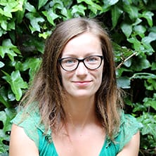 A woman with glasses and light brown hair wearing a green top sits in front of dense green foliage, looking at the camera.