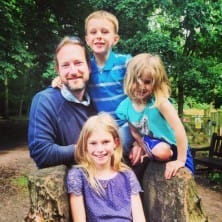 A man sits on a tree stump outdoors with three children, two girls and one boy, all smiling at the camera; trees and a park are in the background.