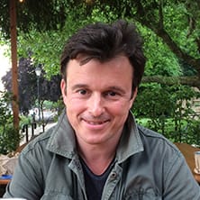 A man with dark hair sits outdoors at a wooden table, smiling, with trees and greenery in the background.