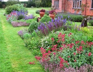 A well-maintained Canada Garden with vibrant red, purple, and green plants borders a neatly mowed lawn next to a brick house with large windows.
