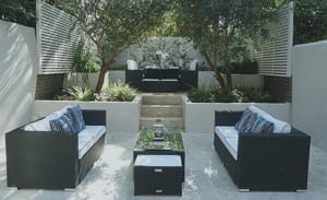Modern outdoor patio with black wicker furniture, white cushions, a glass coffee table, and greenery creates a green room feel around the raised dining area in the background.