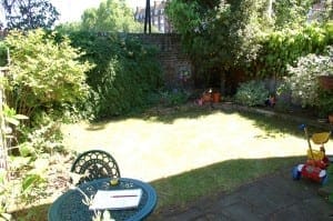 A small chillout garden with green shrubs, a patchy lawn, a round metal table and chair, children’s toys, potted plants, and a brick wall in the background.