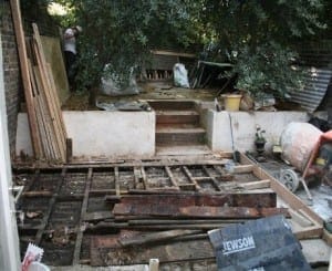 A backyard area under renovation with exposed wooden beams, construction materials, and tools scattered around; two people work near trees in the background, adding touches of Eastern Exotic style to the evolving space.