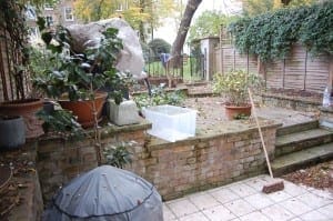 A backyard patio with potted plants, a clear plastic storage bin, a broom, and a covered object, bordered by a wooden fence and greenery, creates an inviting green room atmosphere outdoors.