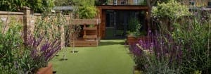 A modern backyard garden with artificial grass, wooden planters, purple and green plants, and a wooden garden shed at the back.