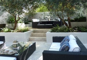 Contemporary outdoor patio with black wicker furniture, white cushions, a glass coffee table, and lush greenery on stepped, white stone terraces.
