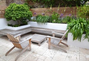 A modern outdoor patio with two wooden chairs, a low table, a built-in corner bench, and raised plant beds filled with green foliage and flowers.