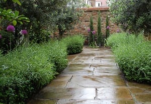 A wet stone pathway winds through lush front gardens adorned with green shrubs, purple flowers, and trees, leading to a brick wall framed by tall, narrow plants.