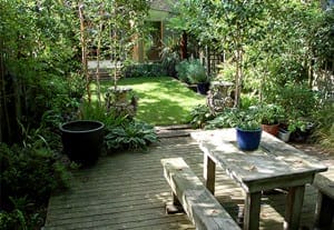 Wooden patio with a rustic table and benches, surrounded by potted plants, leading to a grassy Family Gardens area filled with trees and shrubs.