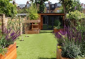 This landscaped garden features artificial grass, wooden planters filled with purple flowers and shrubs, and a wooden shed at the back, set against residential buildings in the background.