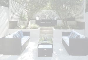 Modern outdoor patio in a garden home with black wicker sofas, white cushions, a glass coffee table, and decorative pillows, surrounded by lush greenery and white privacy panels.