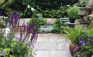 A sketch of a small patio garden with stone paving, purple and white flowers, green plants, a metal chair, a round table, and a stone birdbath set against a charming brick wall.