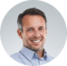 A man with short brown hair and a beard, wearing a light blue collared shirt, smiles at the camera against a plain light background.