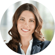 A woman with long brown hair, wearing a dark blazer and white shirt, smiles at the camera in a bright indoor setting.