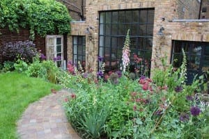 A brick path curves through a lush cottage garden filled with flowering plants, leading to a brick building with large glass windows and ivy climbing the walls.