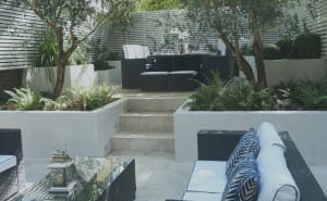 Modern outdoor patio with symmetrical white walls, black wicker furniture, blue and white cushions, potted plants, and a dining area elevated by stone steps.