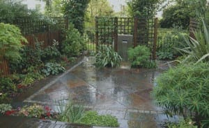 A small garden patio with wet slate stone tiles, surrounded by lush green plants and wooden trellises, featuring a modern Slate & Steel vertical water fountain.