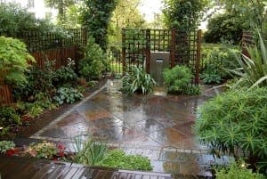 A wet, paved garden patio with lush green plants, trees, flower beds, Slate & Steel elements, wooden fencing, and a water feature in the background.