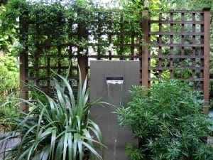 A modern garden water feature with a rectangular slate & steel fountain, surrounded by green plants and wooden lattice panels in the background.