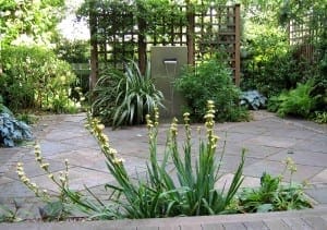 A neatly paved garden patio with a central Slate & Steel water feature, surrounded by green plants, trellises, and various foliage.