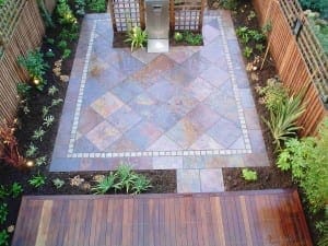 A small, fenced backyard garden with a central square patio of slate tiles and steel accents, surrounded by planting beds and wooden decking in the foreground.