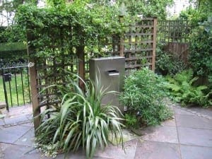 A slate & steel mailbox stands among green plants and wooden lattice panels in a paved garden corner.