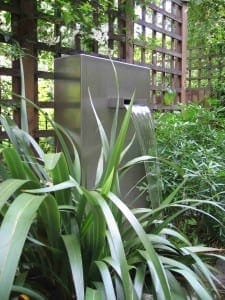 A Slate & Steel fountain with a water spout stands among green foliage and lattice fencing in a garden setting.
