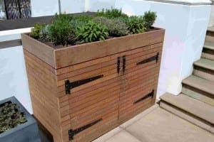 A wooden binsheds storage box with black metal hinges and a planter on top, filled with green herbs, sits next to a set of outdoor stairs.
