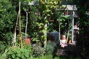 A young child in a colorful dress sits on a swing in lush gardens, surrounded by vibrant plants and blooming flowers.